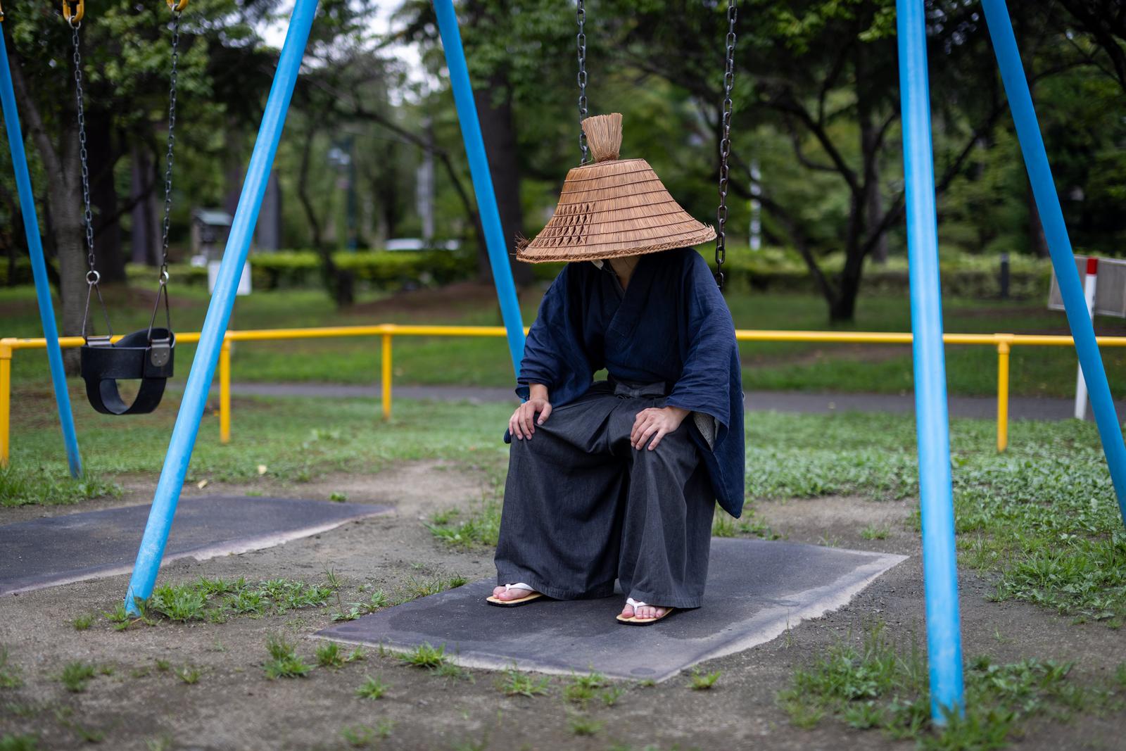 公園のブルーのブランコに座る笠をかぶった侍姿の男性［モデル：まーこ］