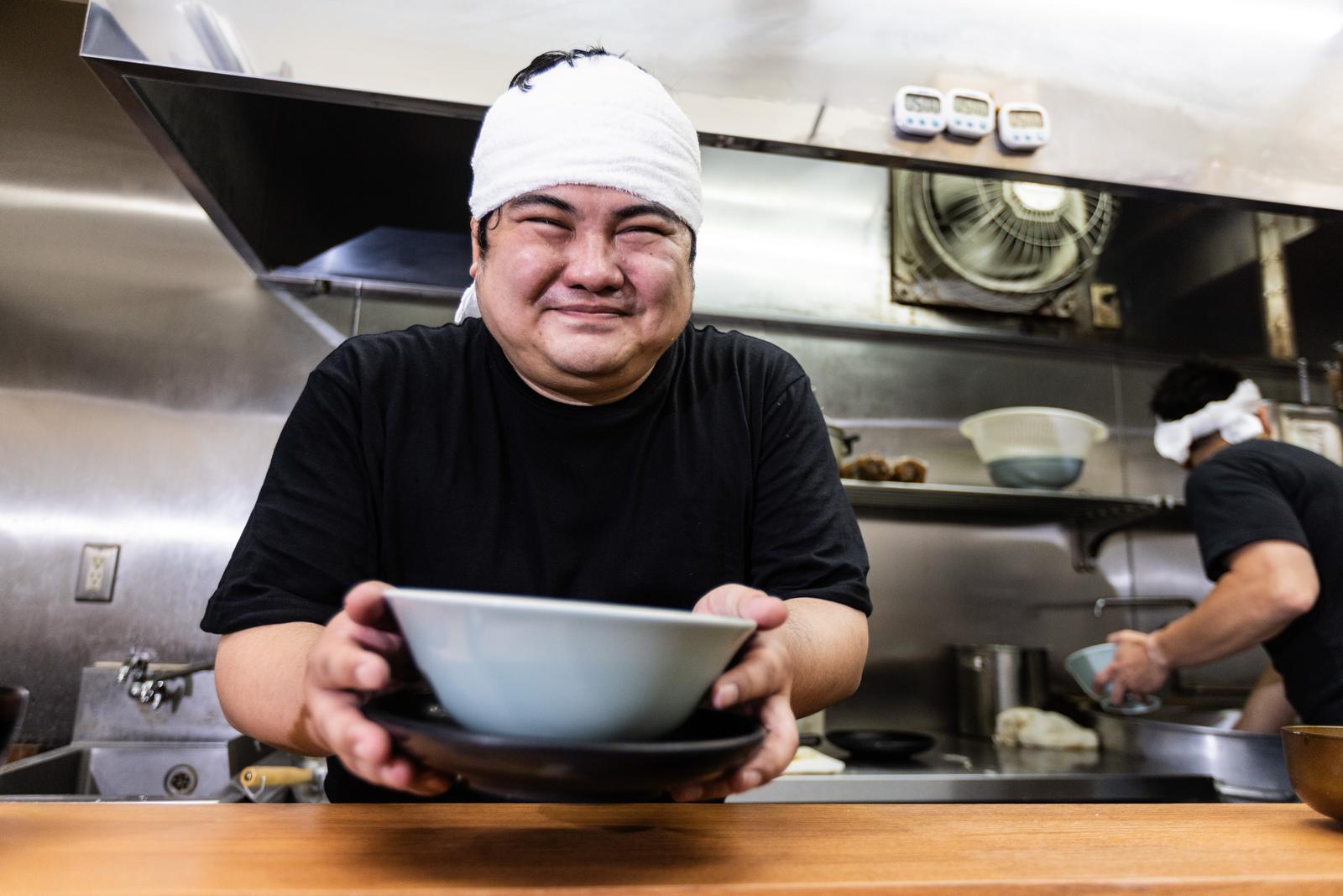 ラーメン店の厨房で丼を持ってほほ笑む店長［モデル：段田隼人］