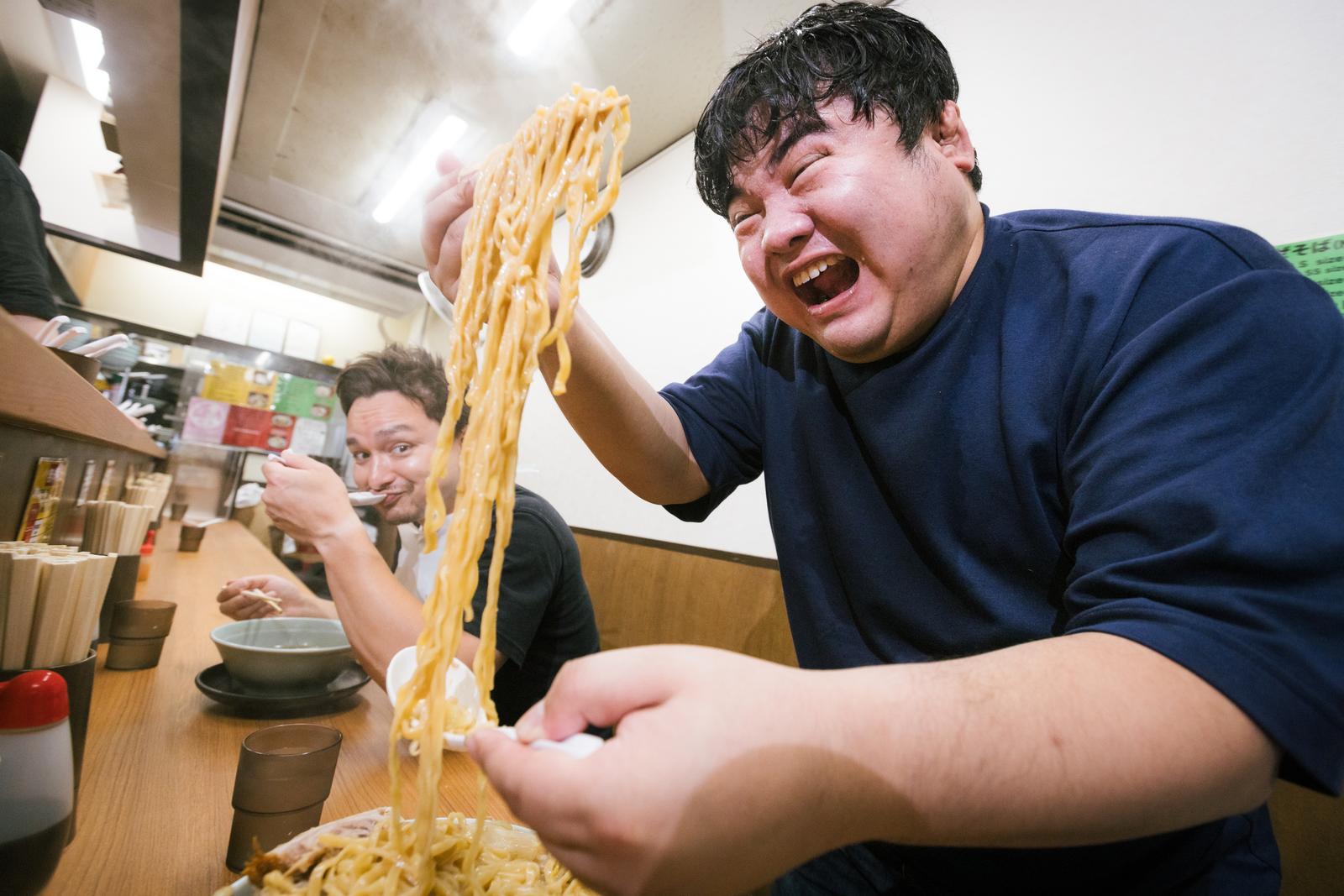 ラーメン店で麺を高く持ち上げて笑う男性［モデル：Max_Ezaki 段田隼人］