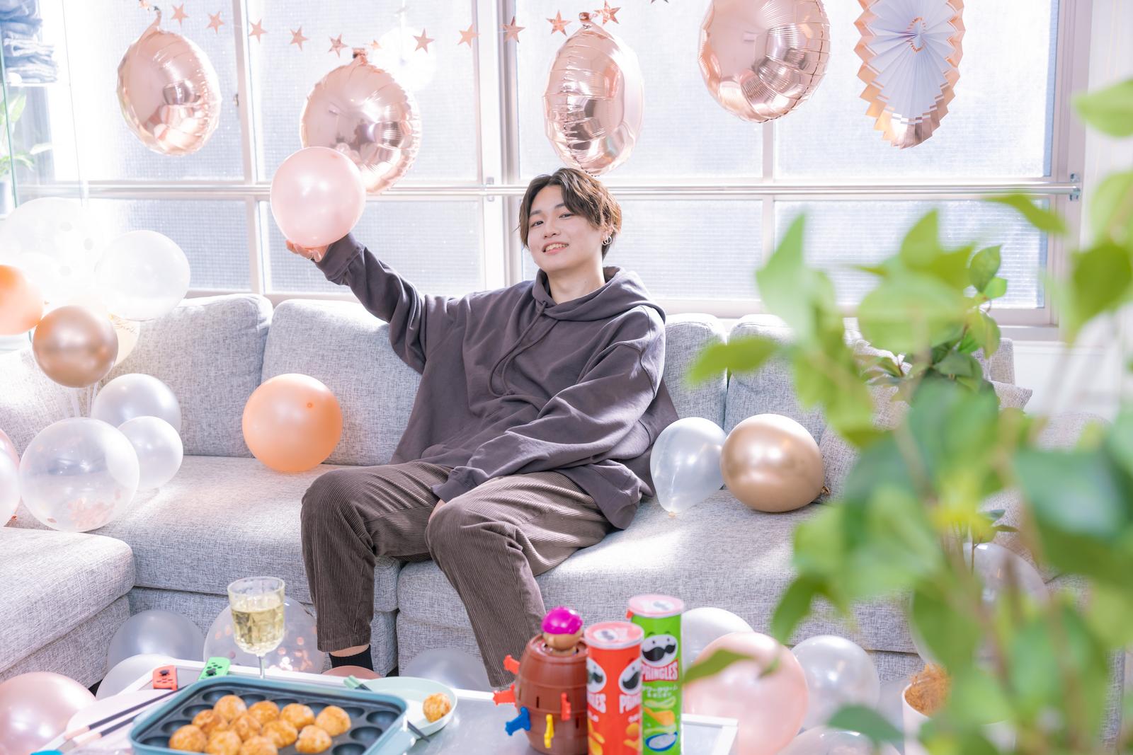ソファーでピンク色の風船を掲げて笑う男性のホームパーティーの様子［モデル：SHUN］