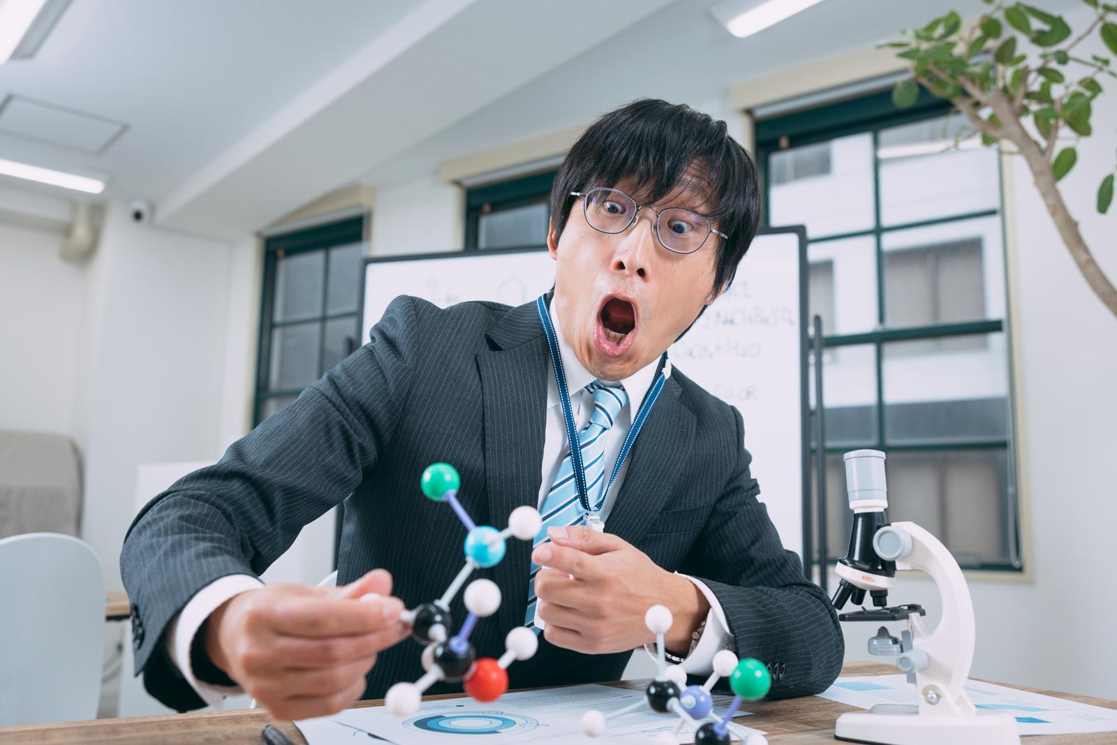 分子構造モデルを持ち驚いた表情の眼鏡をかけた男性研究者［モデル：あづま］