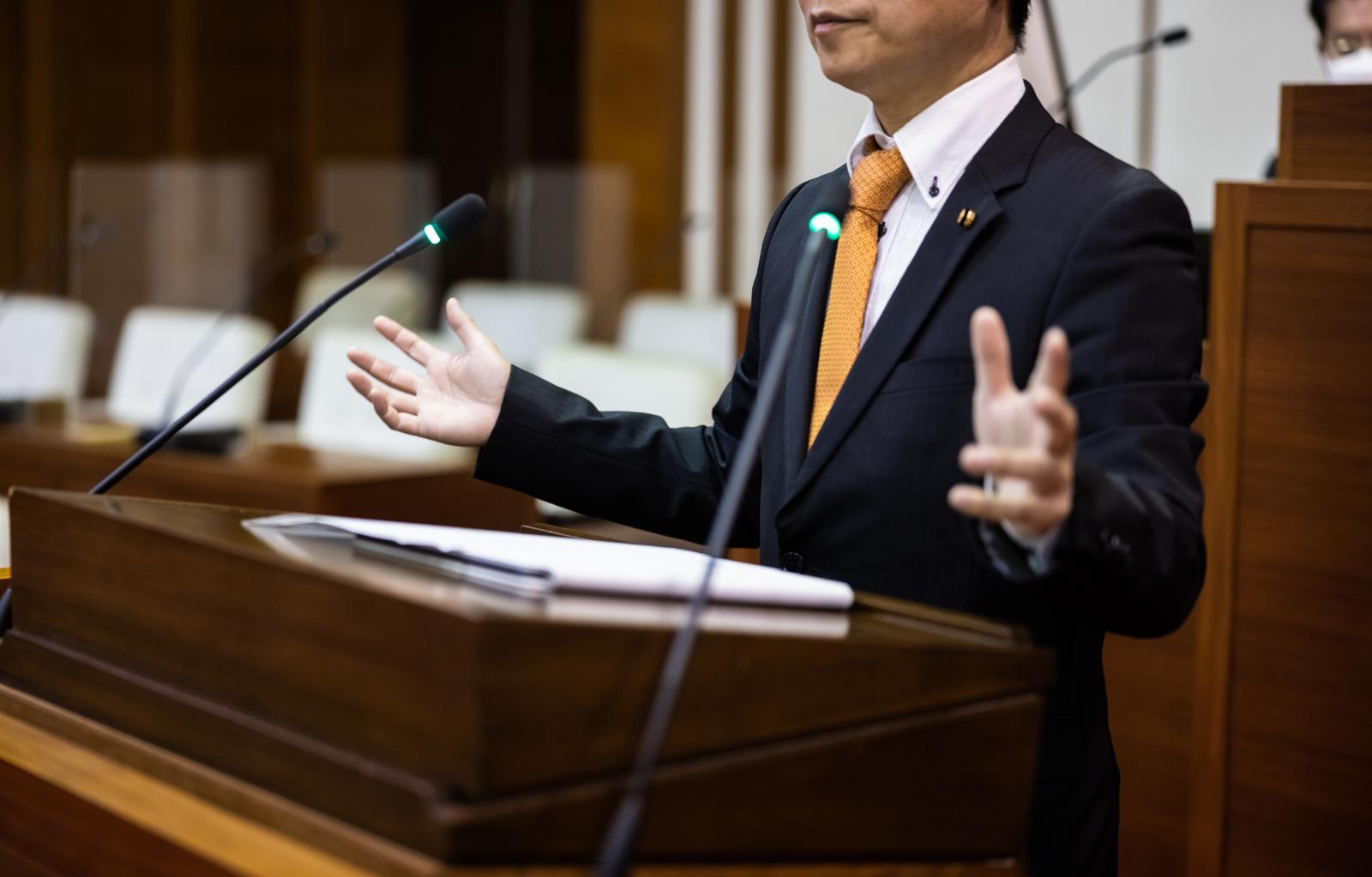 議会で両手を広げて話す紺色スーツの男性議員
