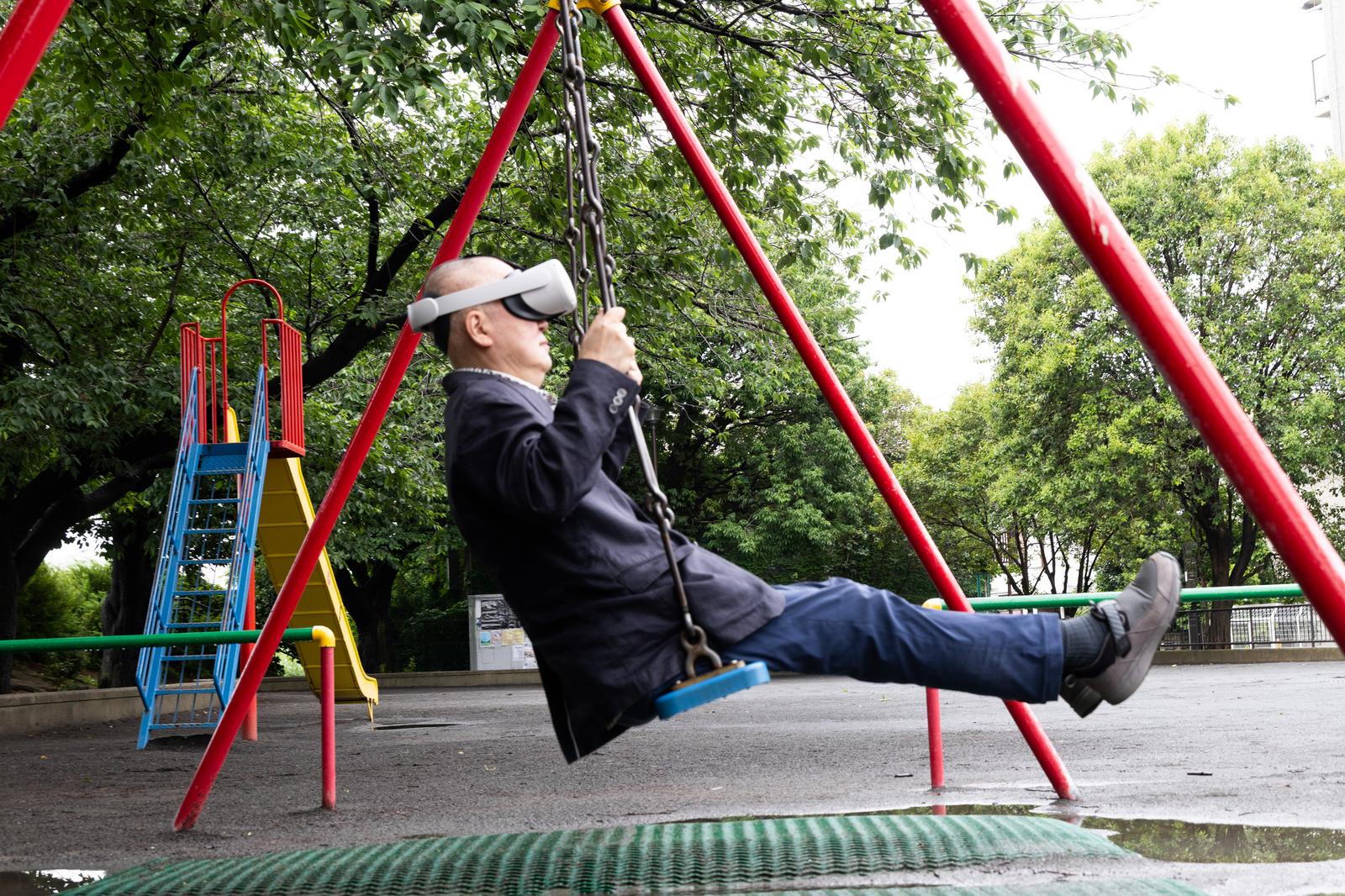 公園でVRゴーグルを付けたスーツ姿の男性がブランコに乗っている［モデル：渡辺カンジ］
