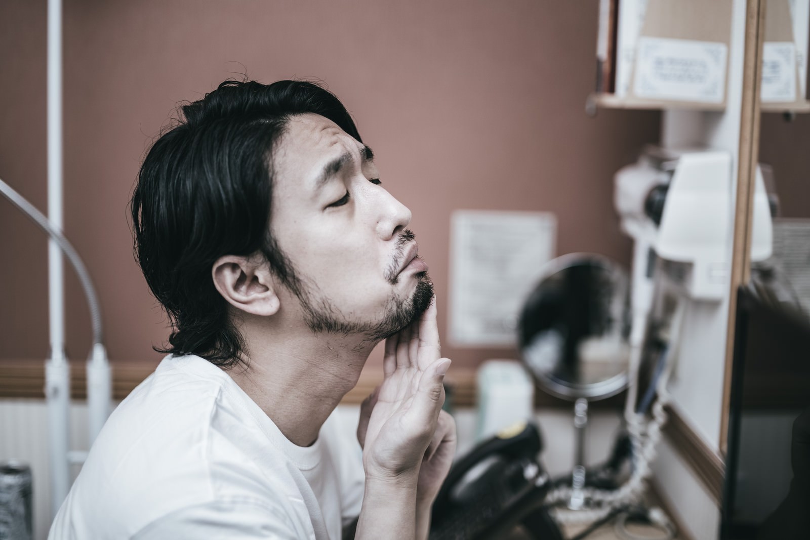 洗面所で無精髭を気にしながら顔に手を当てる男性［モデル：大川竜弥］