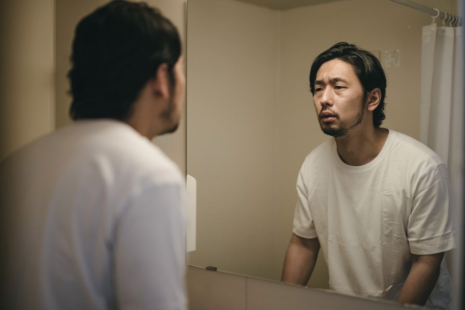 洗面所の鏡の前に立つ無精髭を生やした白いTシャツの男性［モデル：大川竜弥］