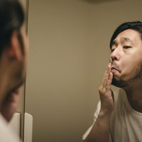 洗面所の鏡で髭の伸び具合を確認する髭面の男性の写真