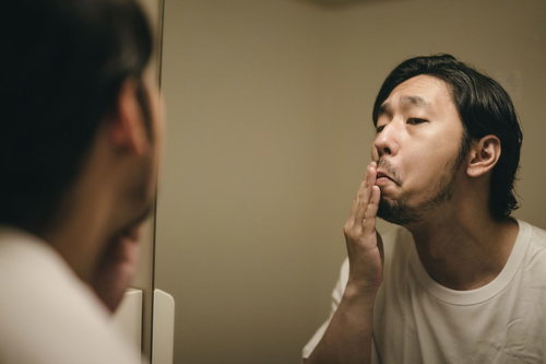 洗面所の鏡で髭の伸び具合を確認する髭面の男性
