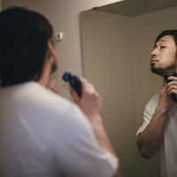 洗面所の鏡の前で電動シェーバーで伸びた髭を剃る男性の写真