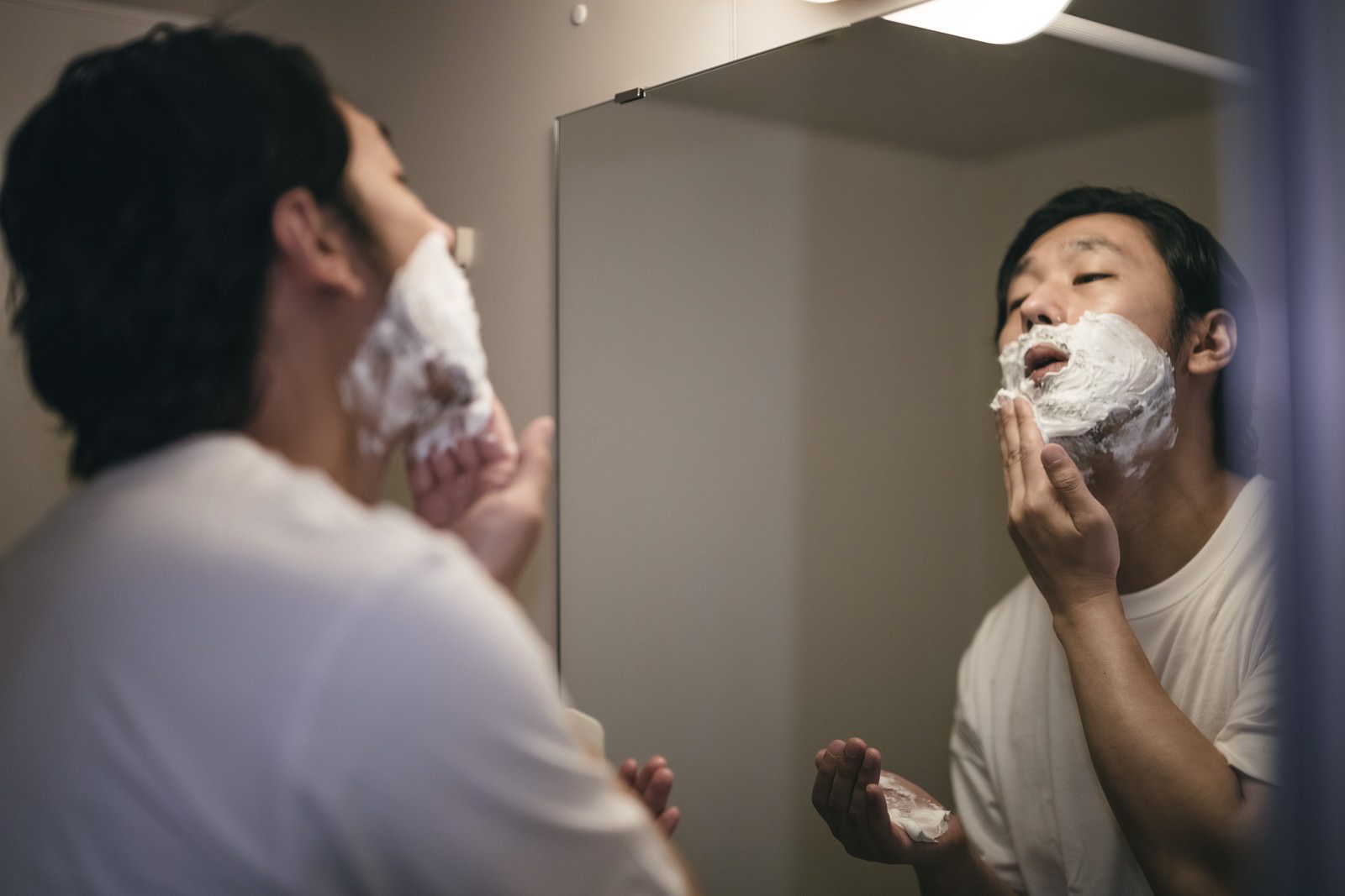 洗面所の鏡の前で髭剃り用の泡を顔に塗る男性［モデル：大川竜弥］