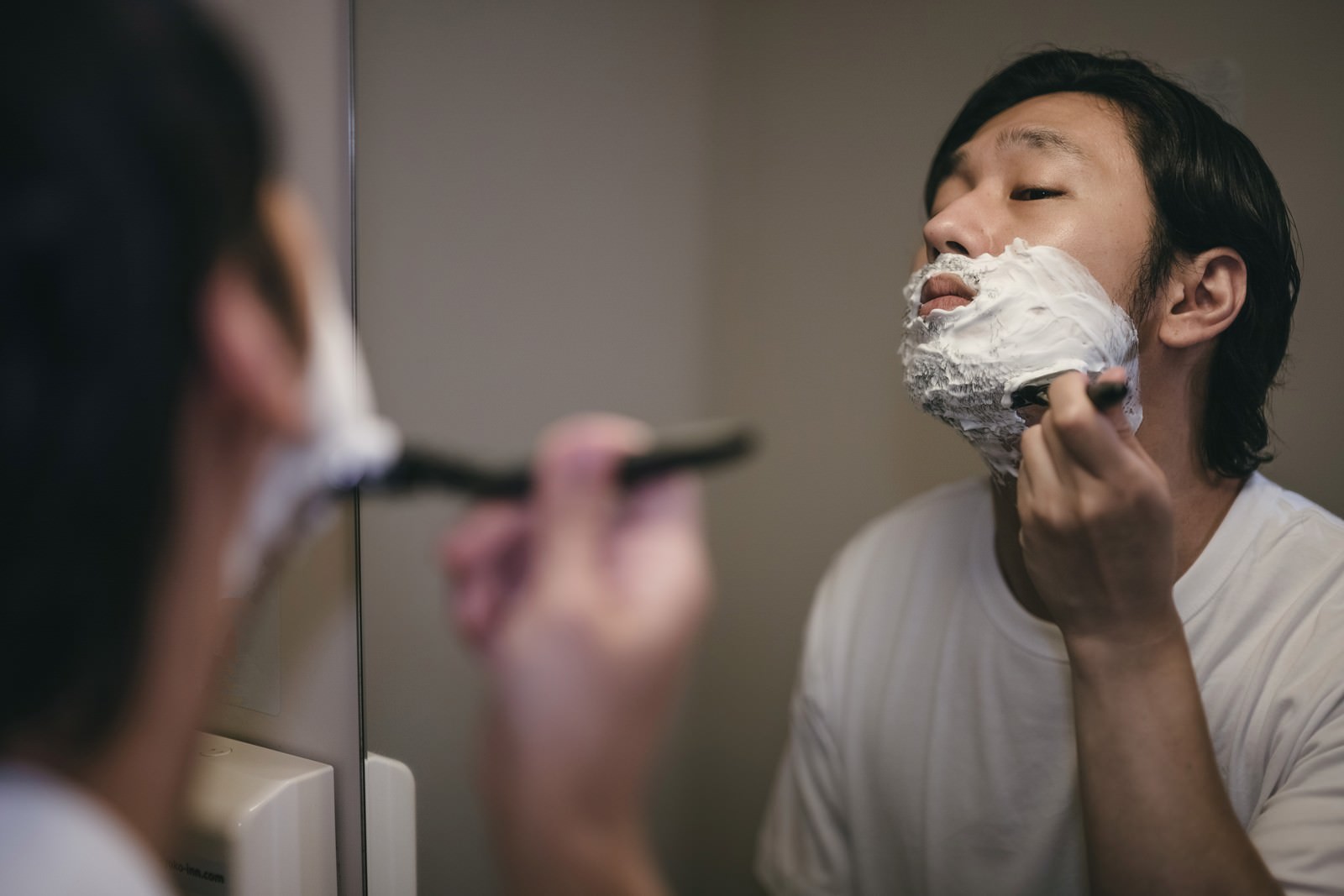 洗面台の鏡の前でシェービングクリームを顔に塗り髭剃りをする男性［モデル：大川竜弥］