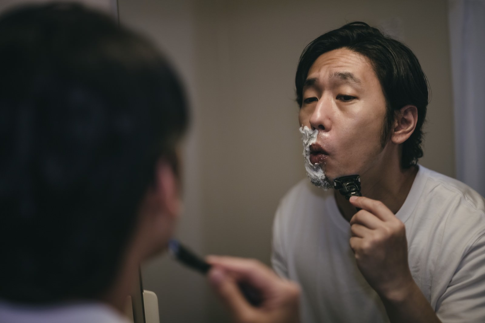 鏡の前で電気シェーバーを頬に当ててひげを剃っている男性［モデル：大川竜弥］