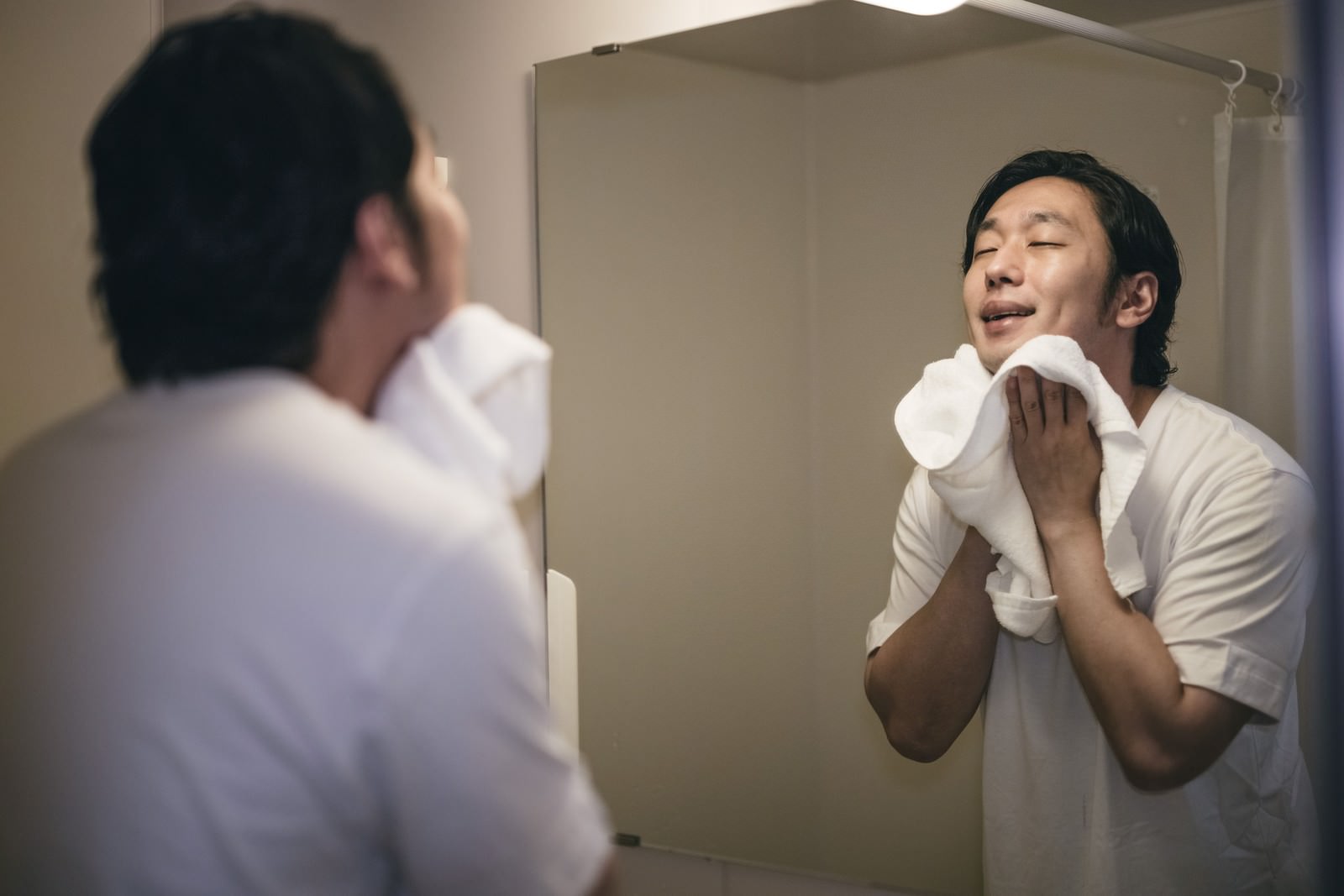 洗顔後に白いタオルを手に持ち、鏡の前でさっぱりとした表情を見せる男性［モデル：大川竜弥］