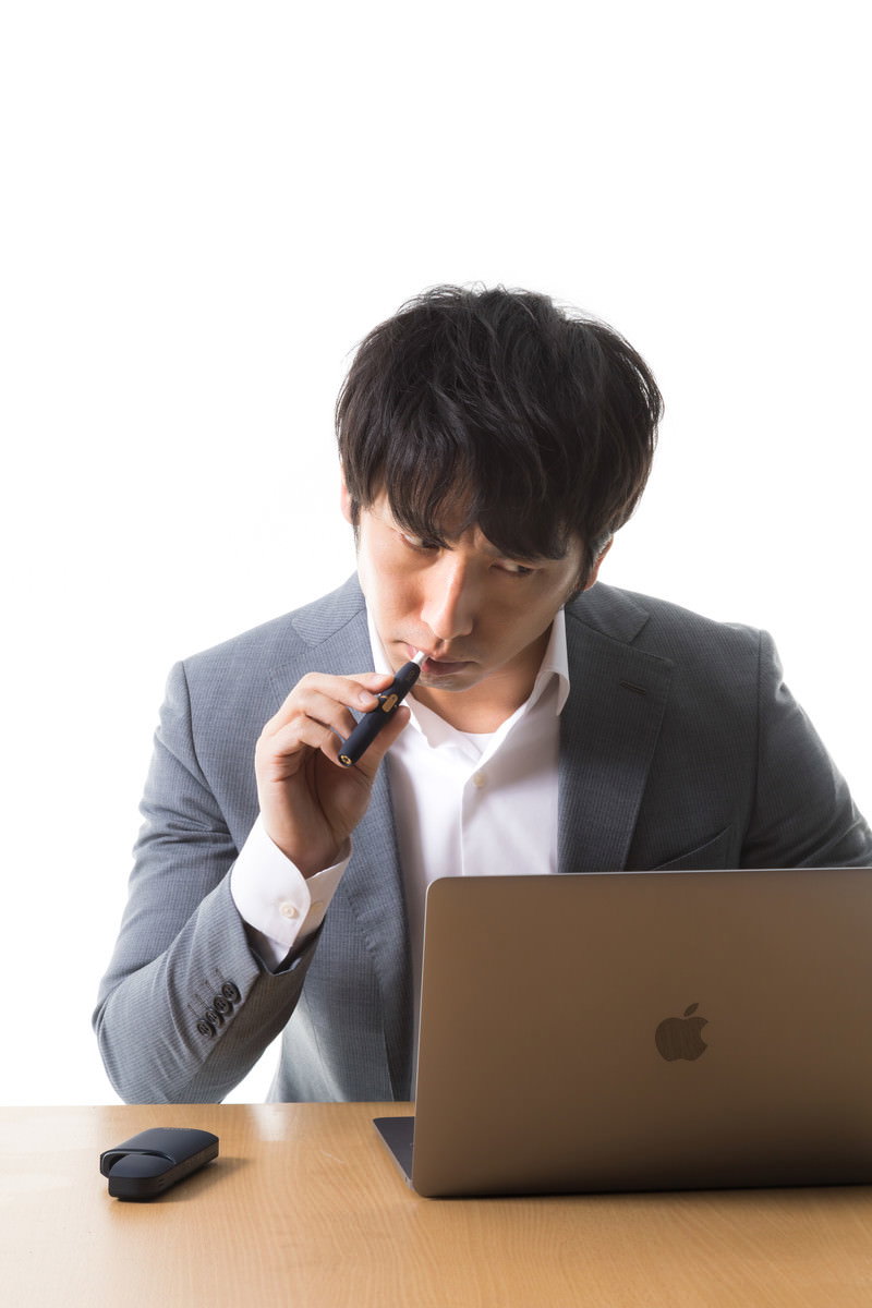 デスクでノートパソコンを使いながら電子タバコを吸うスーツ姿の男性［モデル：大川竜弥］