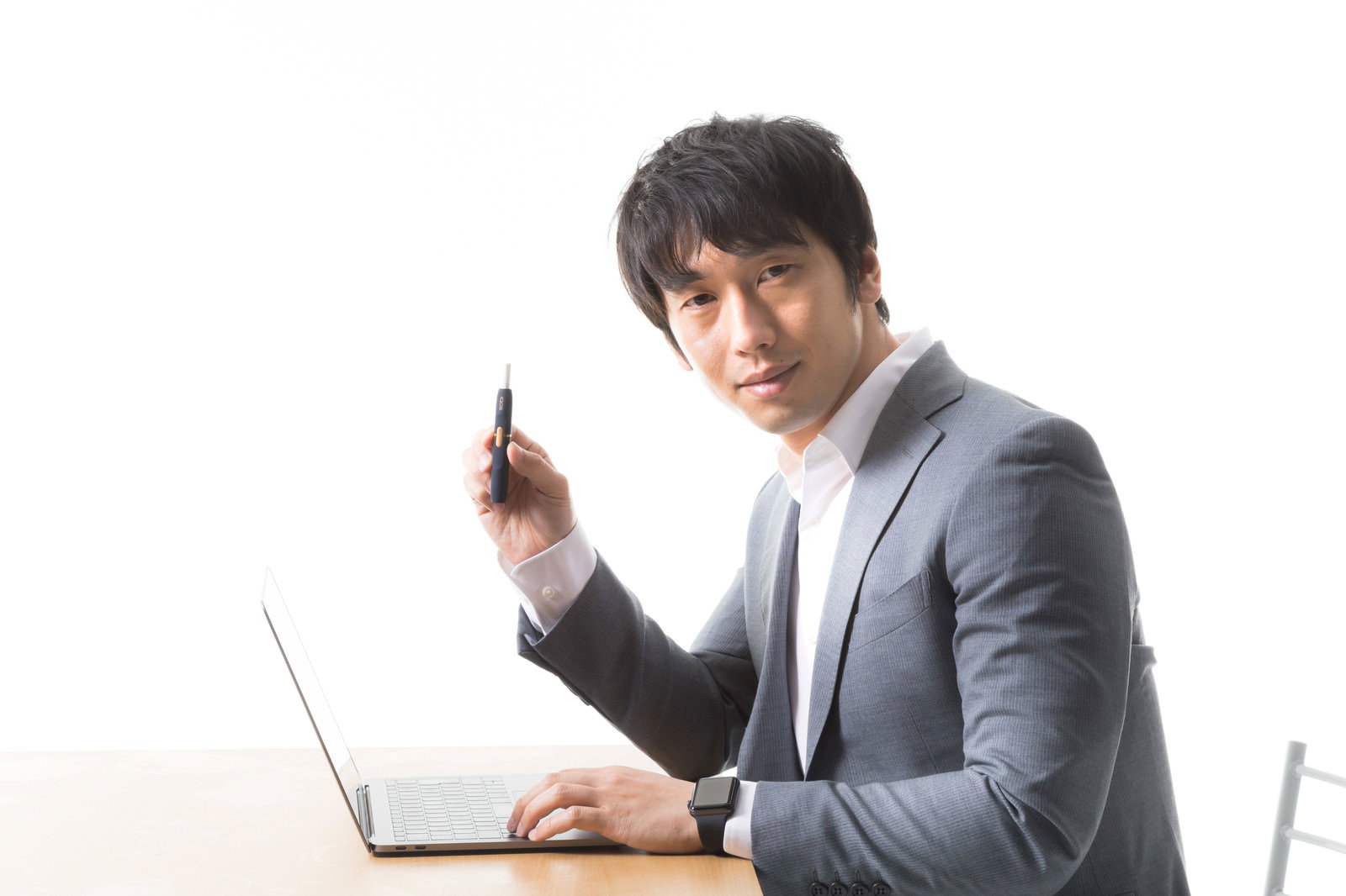グレーのスーツを着た男性がノートパソコンの前で加熱式たばこを手に持っている［モデル：大川竜弥］