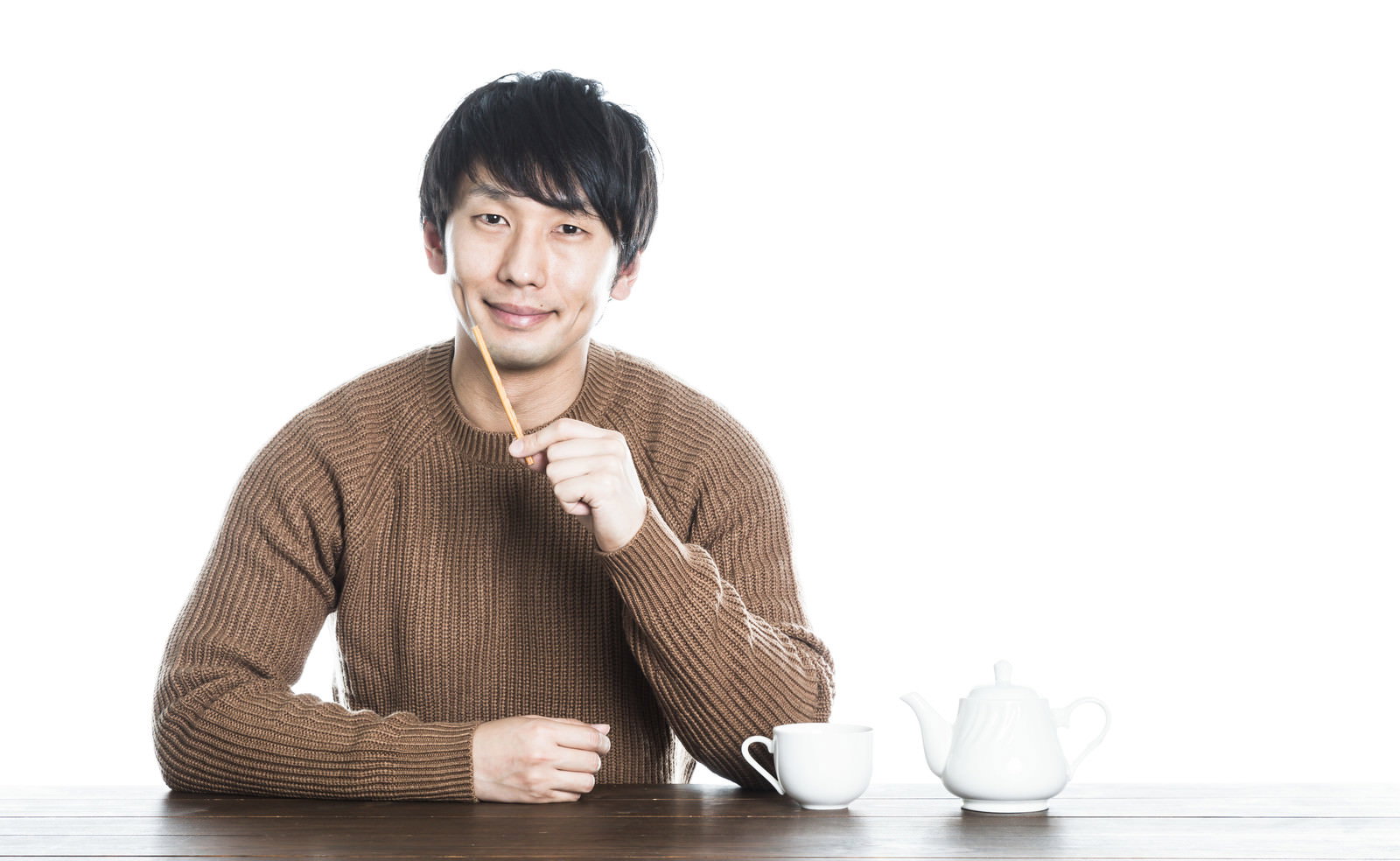 茶色のセーターを着た男性が白いマグカップの前に座り、箸を持ちながら笑顔でくつろいでいる［モデル：大川竜弥］