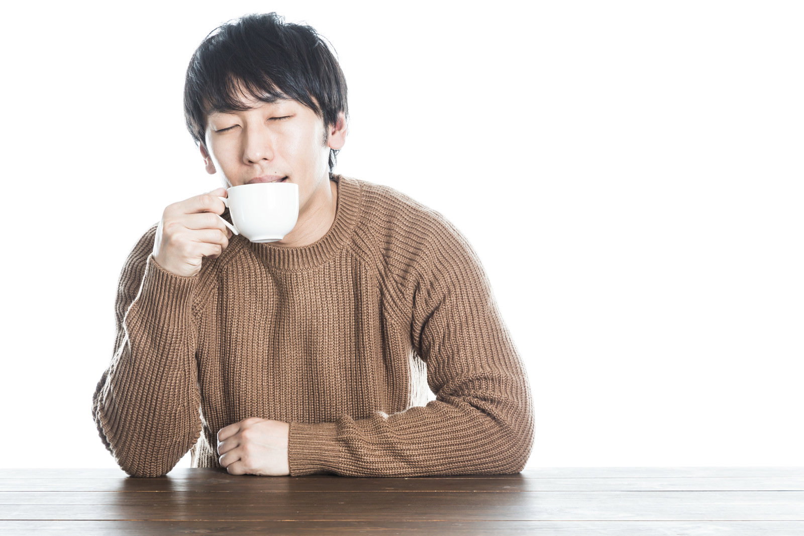 ベージュのニットを着た男性が白いマグカップを両手で持って温かい飲み物を飲んでいる様子［モデル：大川竜弥］