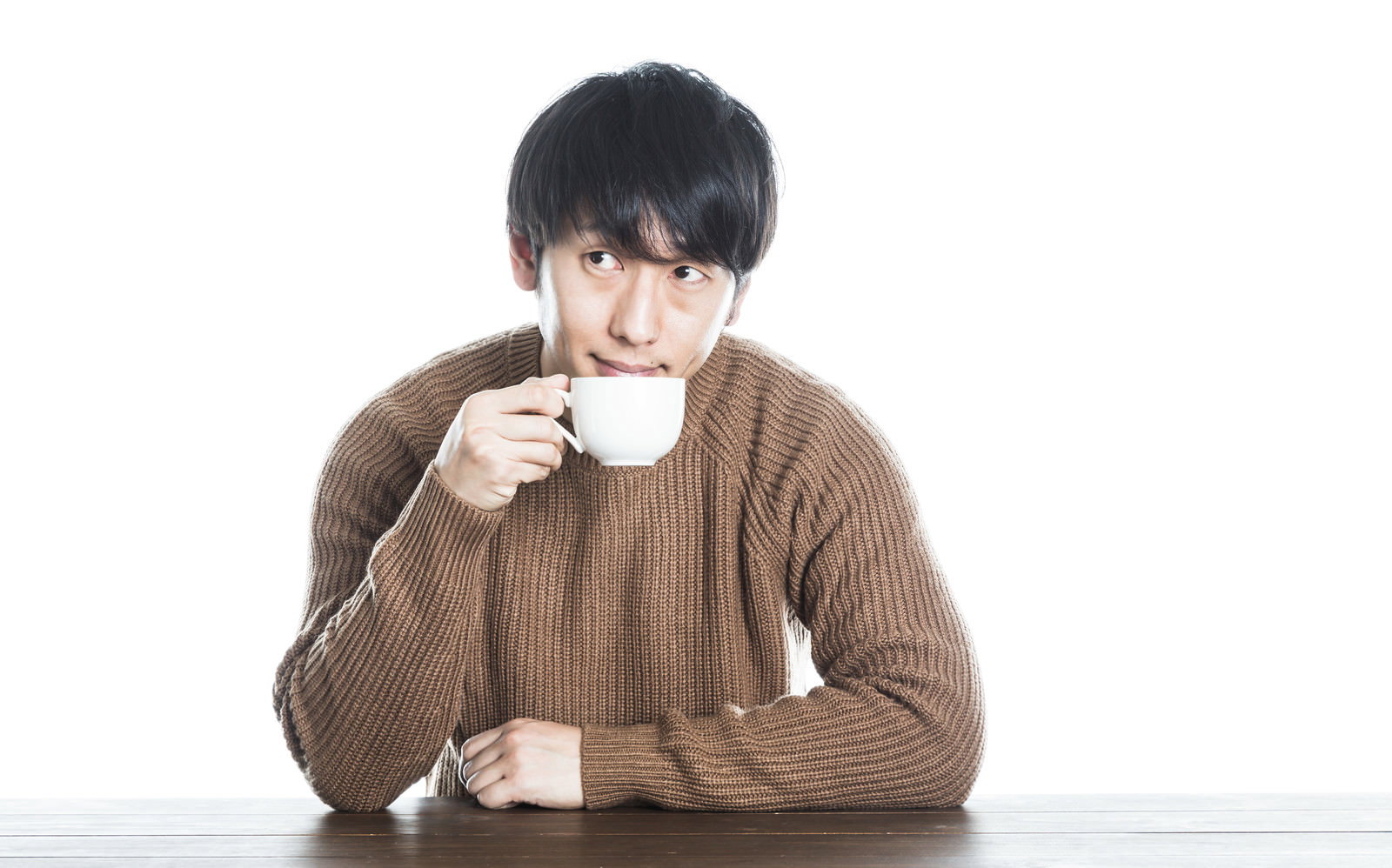 テーブルに向かい白いカップを両手で持つ茶色ニット着用の男性［モデル：大川竜弥］