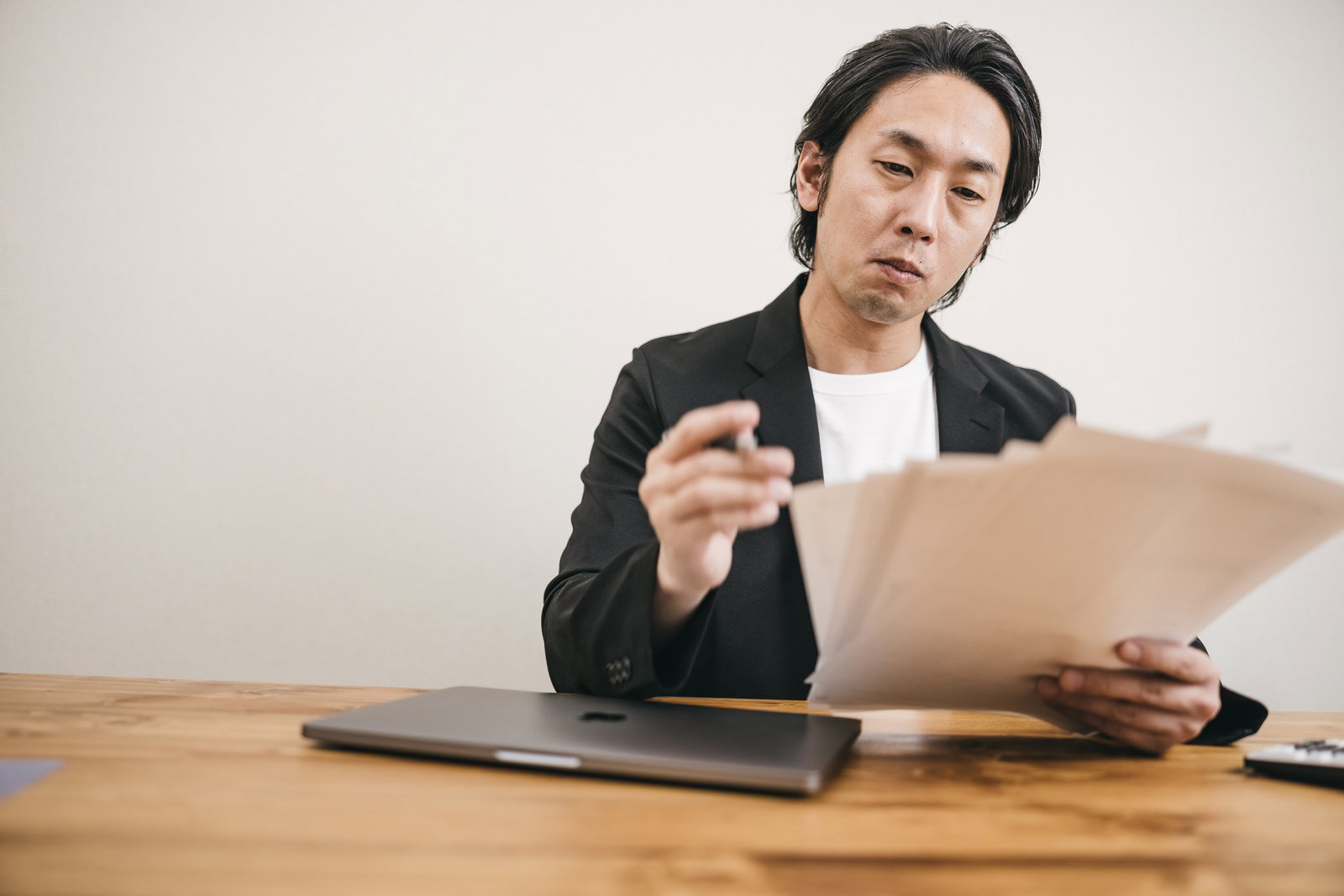 黒いスーツを着た男性が木製テーブルで書類を手に困惑している様子［モデル：大川竜弥］