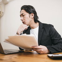 パソコンの画面と手に持つ資料を見比べて考える男性の写真