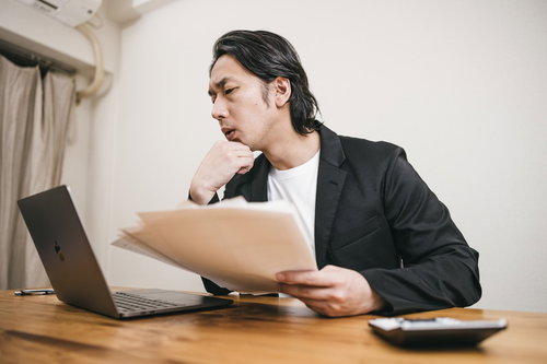 パソコンの画面と手に持つ資料を見比べて考える男性