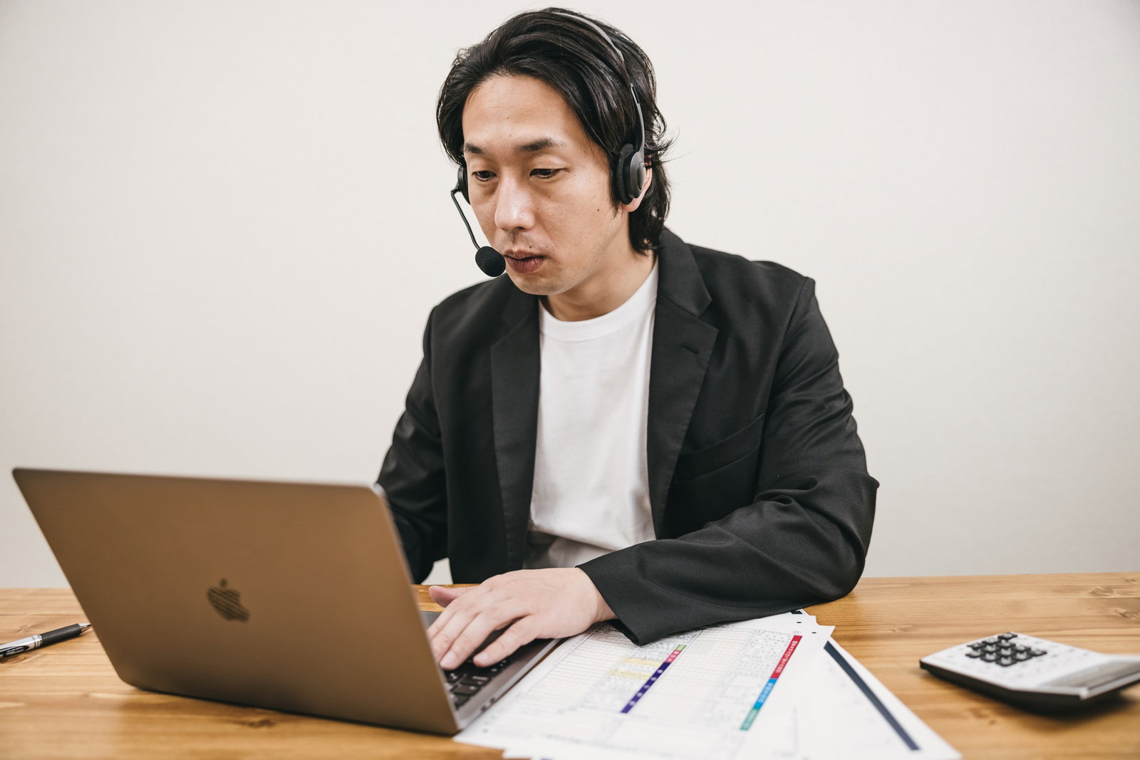 ヘッドセットを着用して木製デスクでノートパソコンを使用する男性［モデル：大川竜弥］