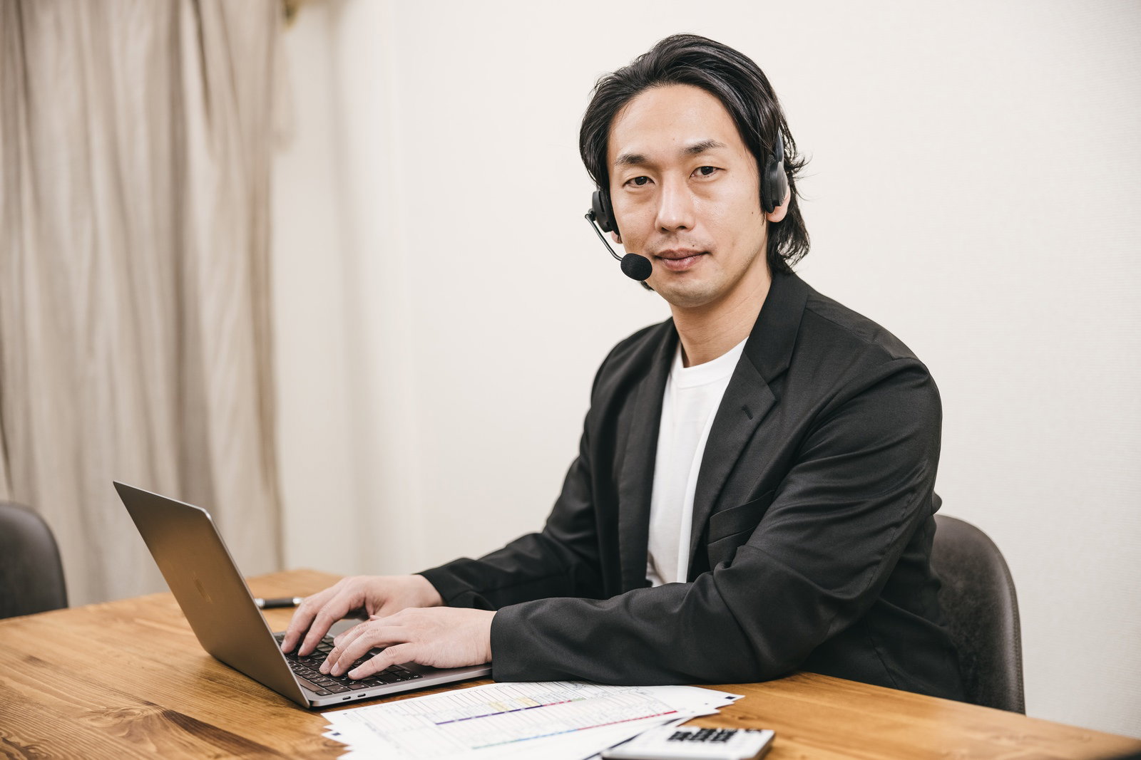 ヘッドセットを着けて木製デスクでパソコン作業をする黒いスーツの男性［モデル：大川竜弥］