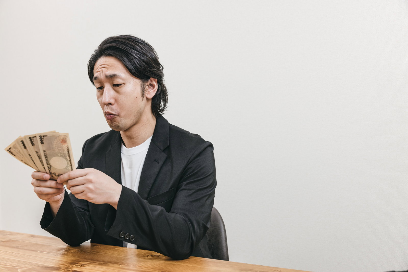 黒いスーツを着た男性が机に座り一万円札の札束を手に持っている［モデル：大川竜弥］