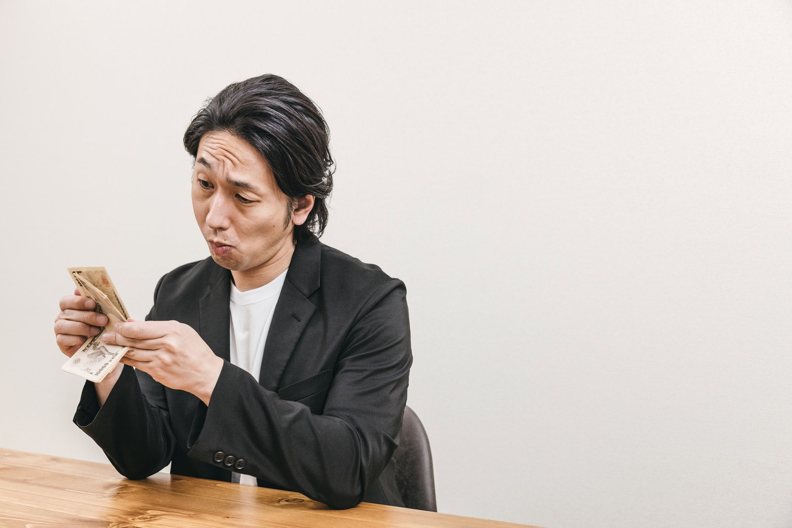 机の上で一万円札の札束を手に取り数える黒いスーツの男性［モデル：大川竜弥］