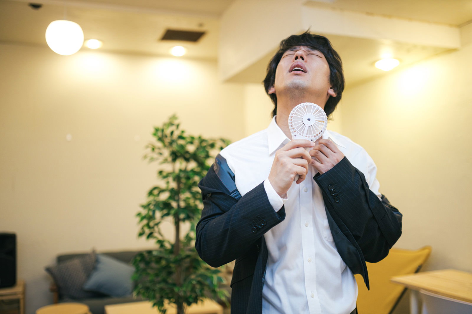 黒いスーツを着た男性が白いハンディ扇風機で首元の汗を乾かしている様子［モデル：大川竜弥］