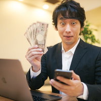 10万円の現金を手に喜ぶビジネスマン、スマートフォン操作の写真