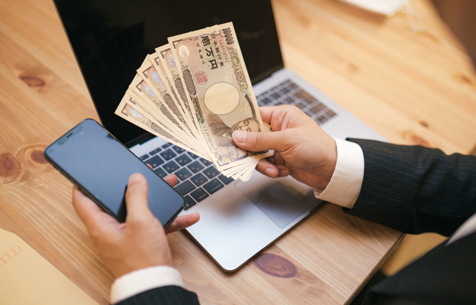 木製テーブルに置かれた万円札の束とスマートフォン、ノートパソコン