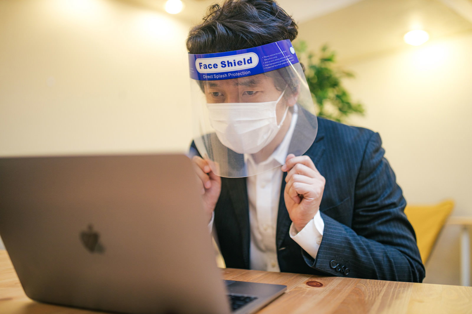 フェイスシールドとマスクを装着してノートパソコンで作業するスーツ姿の男性［モデル：大川竜弥］