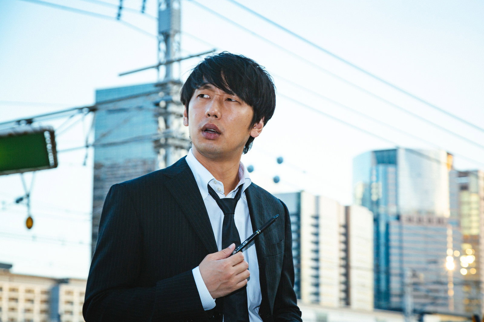 ビル群を背景に黒いスーツを着て困った表情を浮かべる刑事［モデル：大川竜弥］