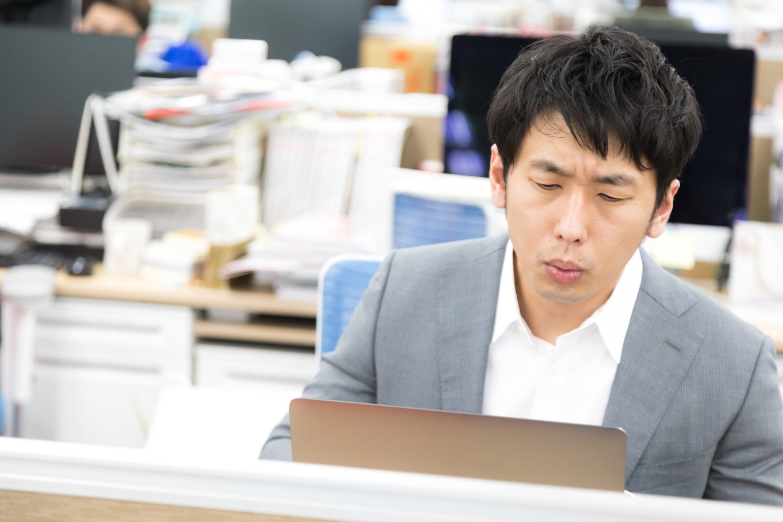 グレーのスーツを着た男性がオフィスのデスクでパソコン画面を見ながら困った表情をしている［モデル：大川竜弥］