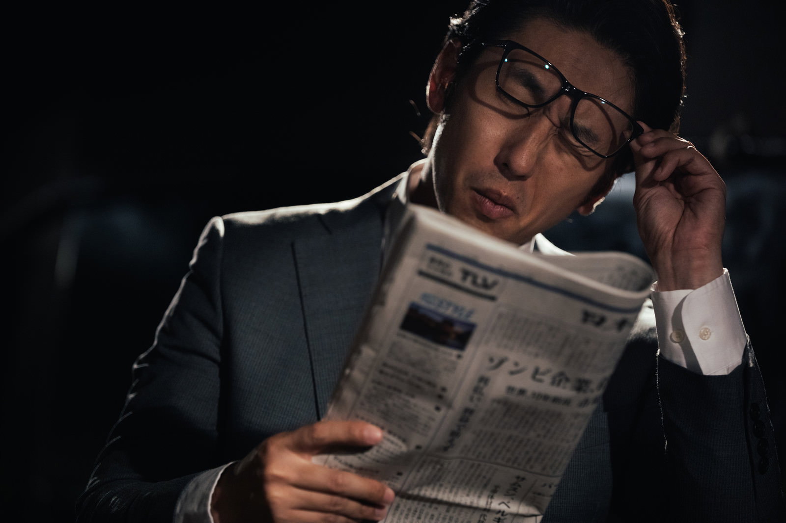 新聞を読みながら眼鏡を押さえて驚いた表情のスーツ姿の男性［モデル：大川竜弥］