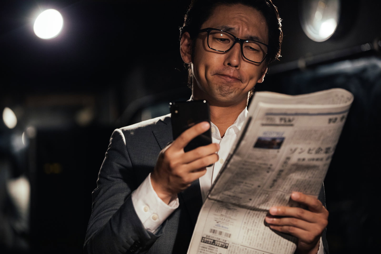 眼鏡をかけたビジネスマンが新聞とスマートフォンを同時に見ている様子［モデル：大川竜弥］