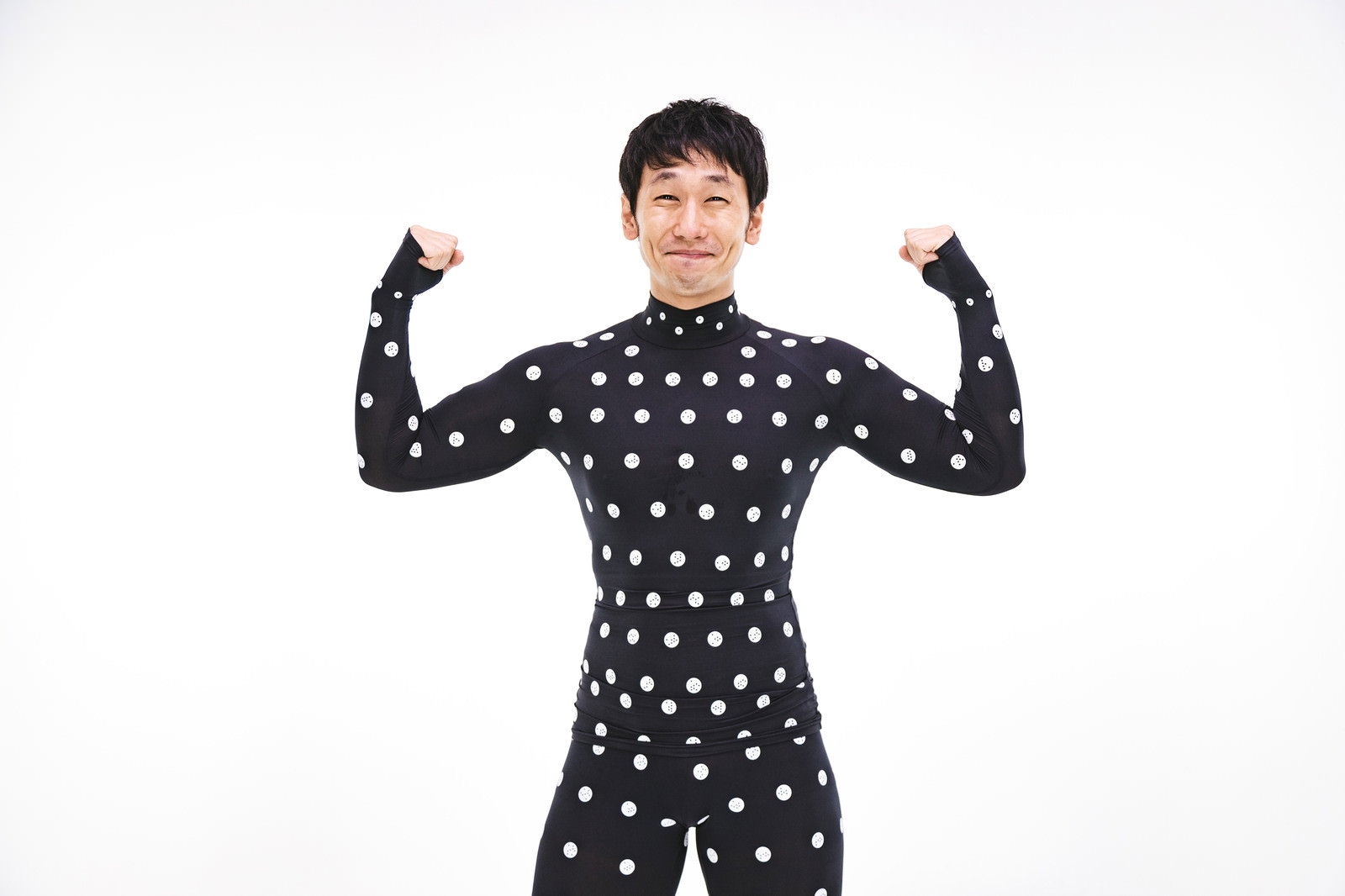 黒いドット柄のボディスーツを着た男性が両腕を上げてポーズをとっている様子［モデル：大川竜弥］