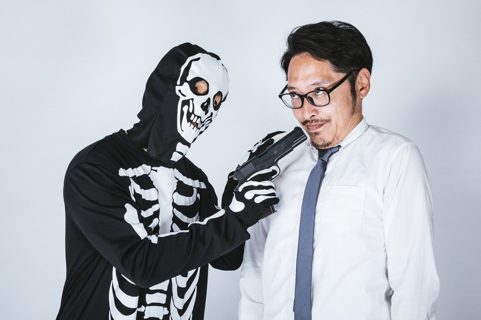 スカルの仮面をつけた黒い服の人物が眼鏡の男性に拳銃を向けている様子［モデル：ゆうせい 大川竜弥］