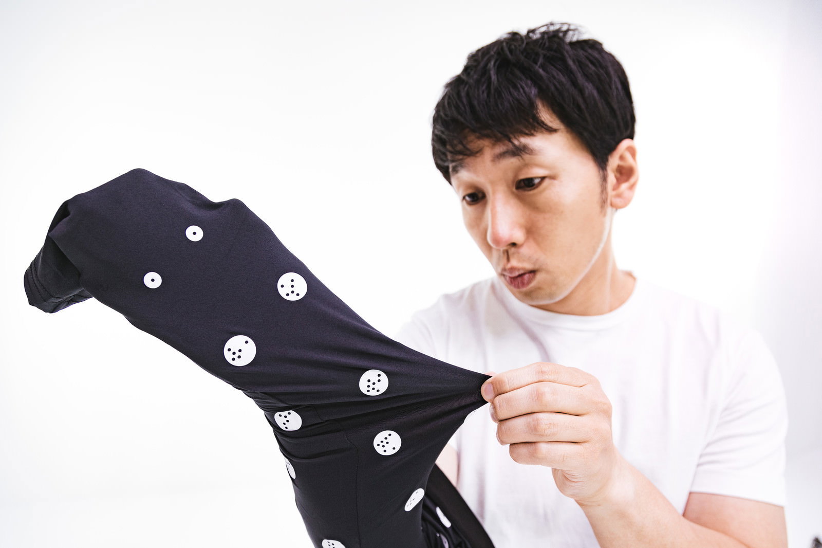 黒色の採寸ボディスーツを手に持ち驚いた表情を見せる白いTシャツの男性［モデル：大川竜弥］
