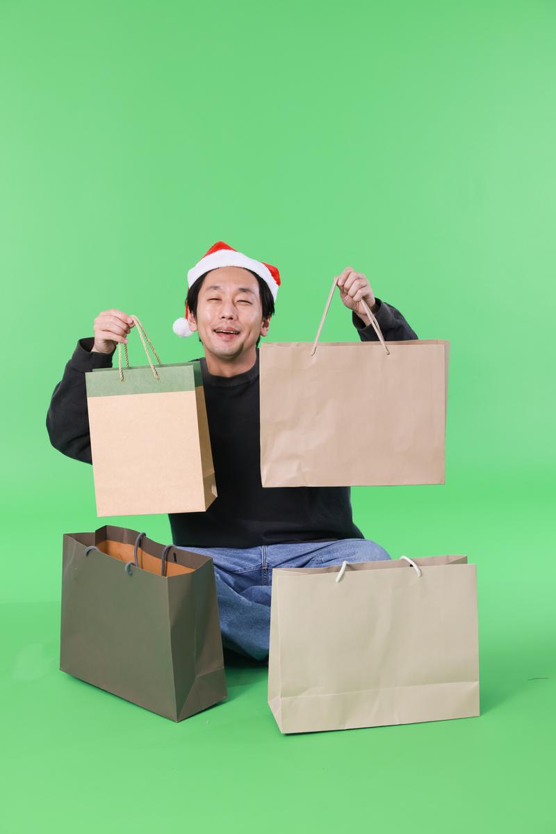 サンタ帽をかぶり複数の買い物袋を持つ男性がクリスマスセールで笑顔［モデル：大川竜弥］