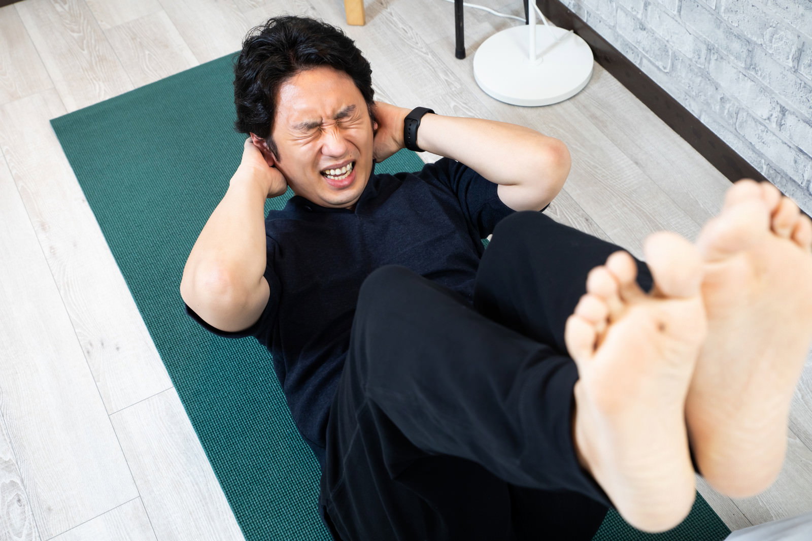 緑のヨガマットで黒いウェアを着た男性が足上げ腹筋をしている［モデル：大川竜弥］