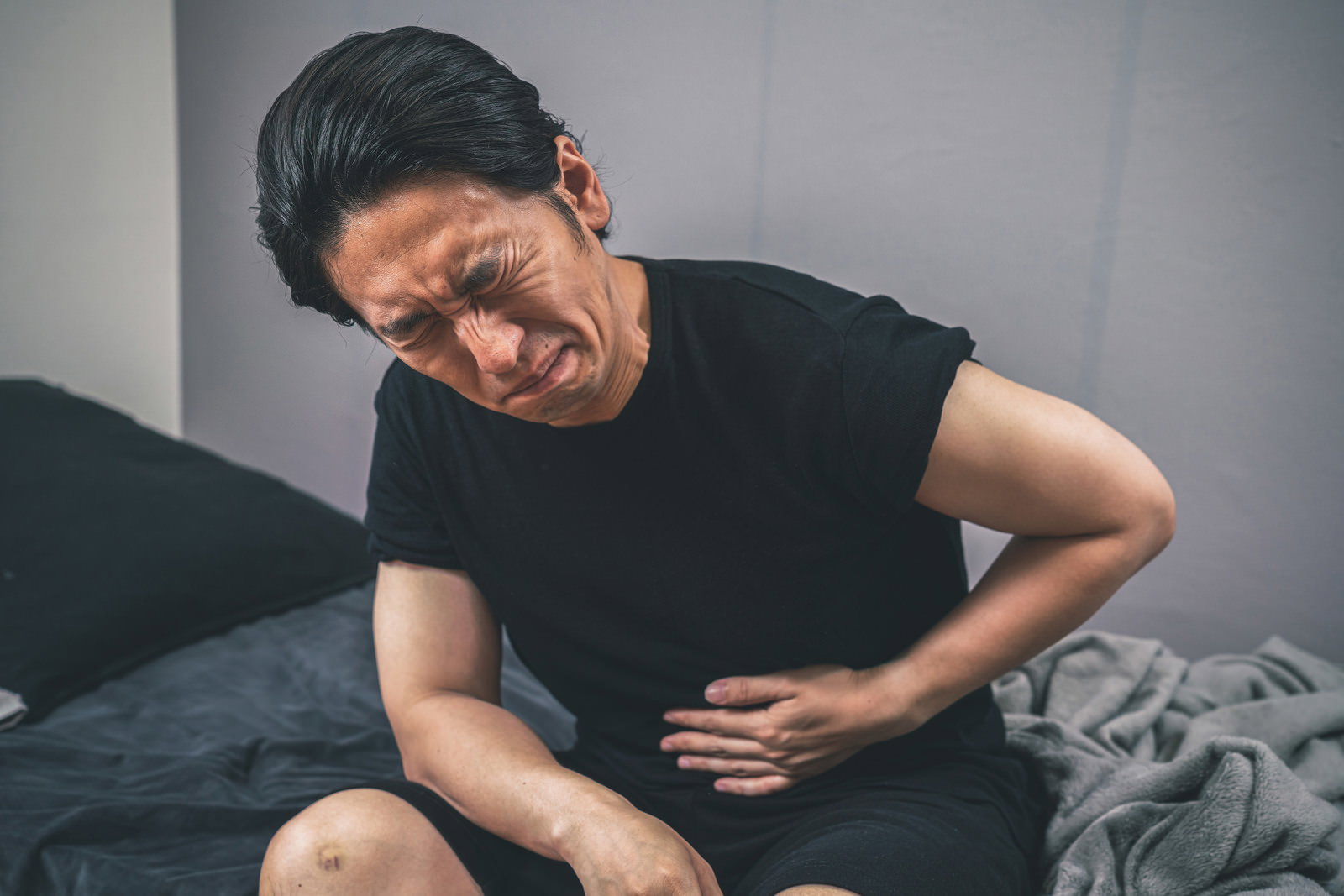 ベッドに座ってお腹を押さえ、腹痛で苦しんでいる男性［モデル：大川竜弥］