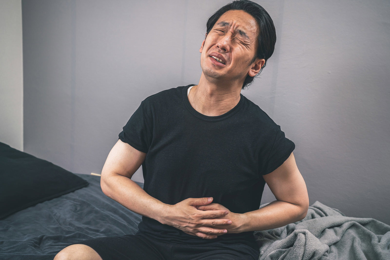 ベッドに座ってお腹を押さえて痛がる黒いTシャツの男性［モデル：大川竜弥］