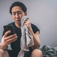 スマホで天気予報を見たら今日も熱帯夜、タオルで汗を拭う男性の写真