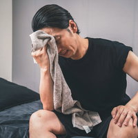 ベッドで寝起きにタオルで汗を拭う男性、熱中症や発熱の体調不良の写真
