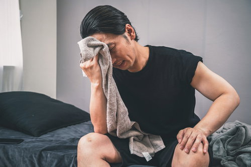 ベッドで寝起きにタオルで汗を拭う男性、熱中症や発熱の体調不良