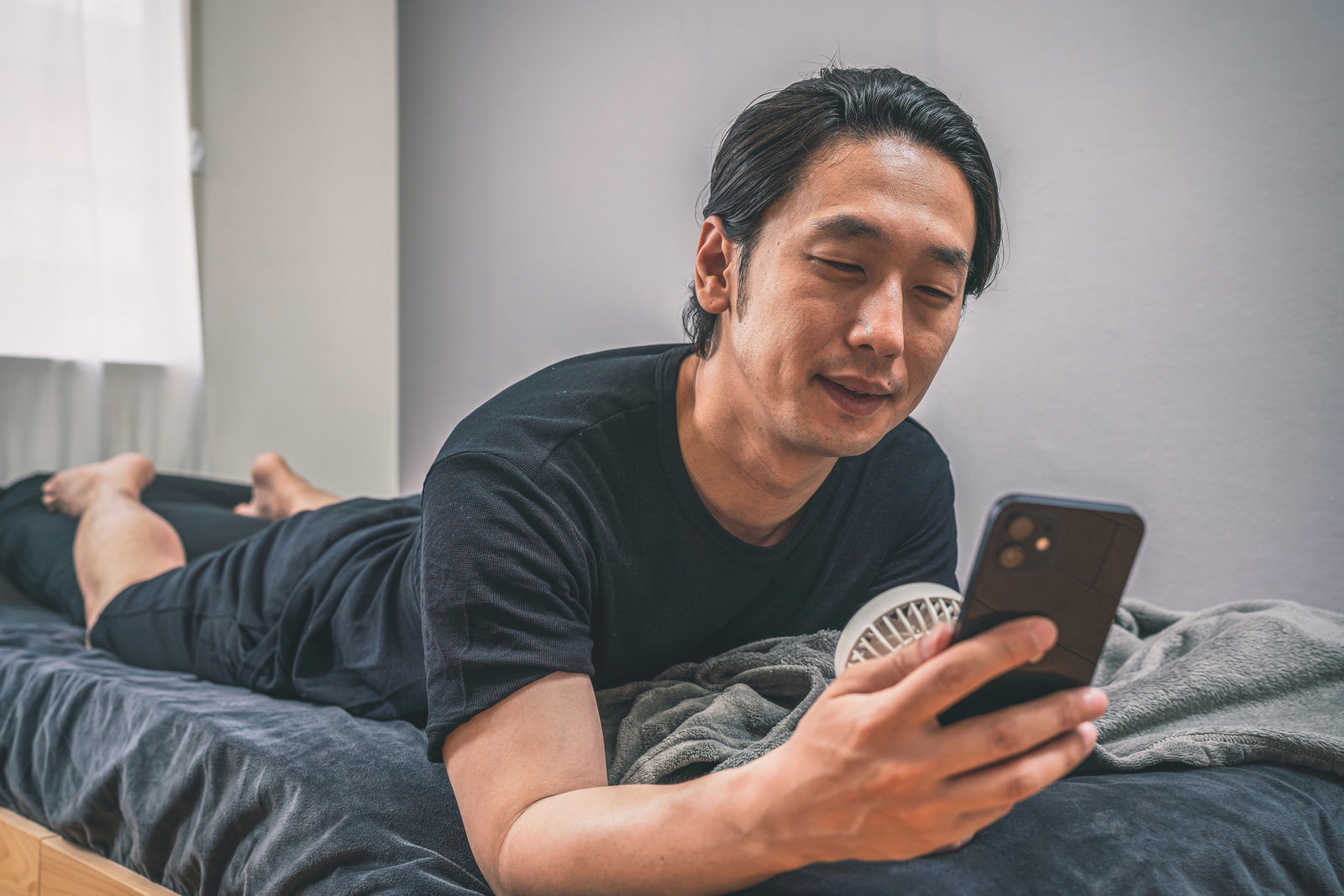 ベッドに横になりながらスマートフォンを操作する男性［モデル：大川竜弥］