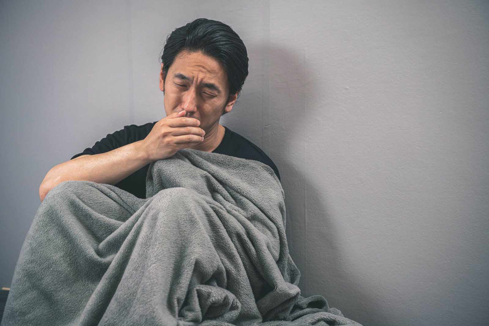 毛布をかぶって壁際に座り咳をする男性［モデル：大川竜弥］