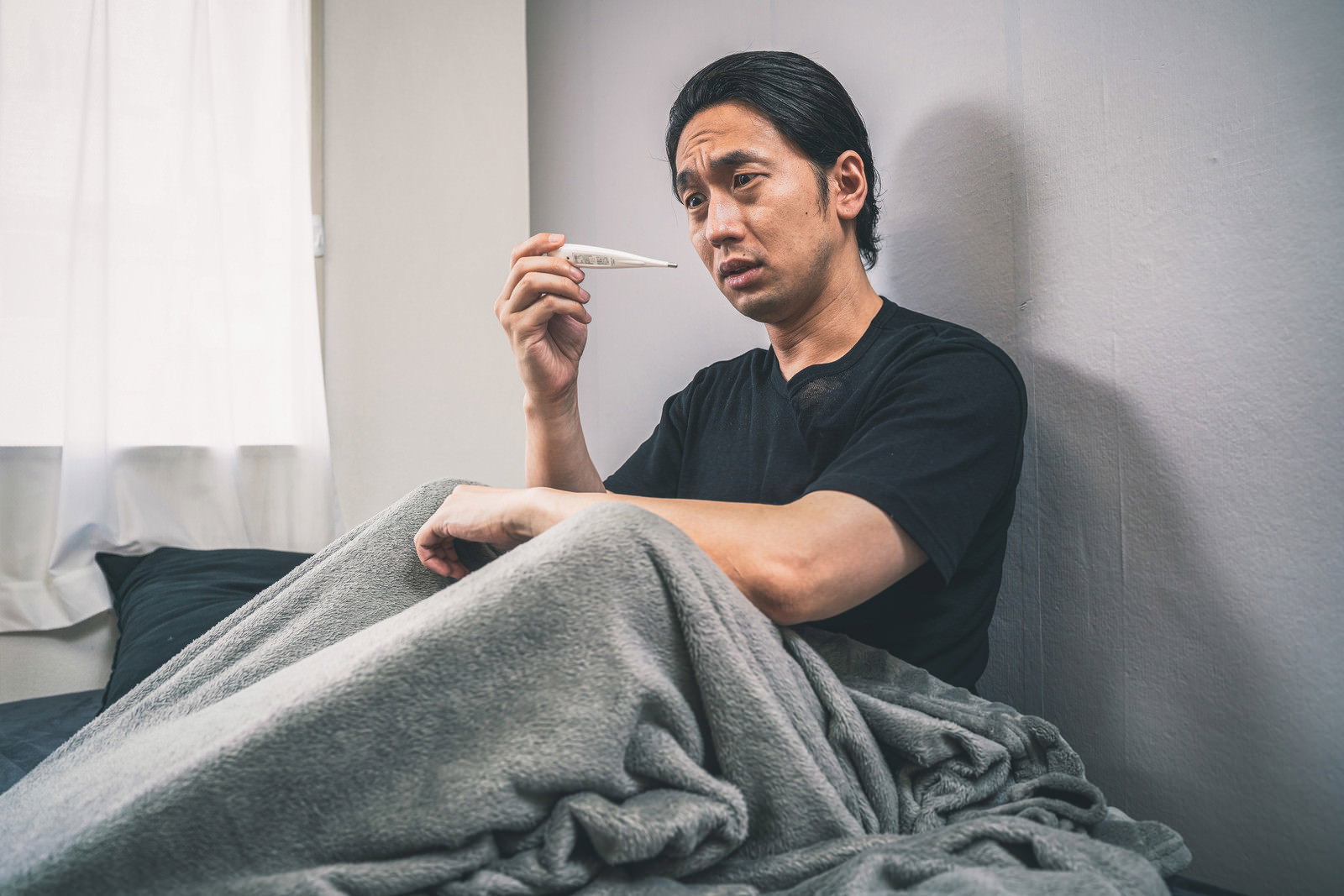 ベッドで体温計を見つめる男性［モデル：大川竜弥］
