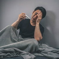 突然の発熱に頭を抱える男性、ベッドで体温計を確認するの写真
